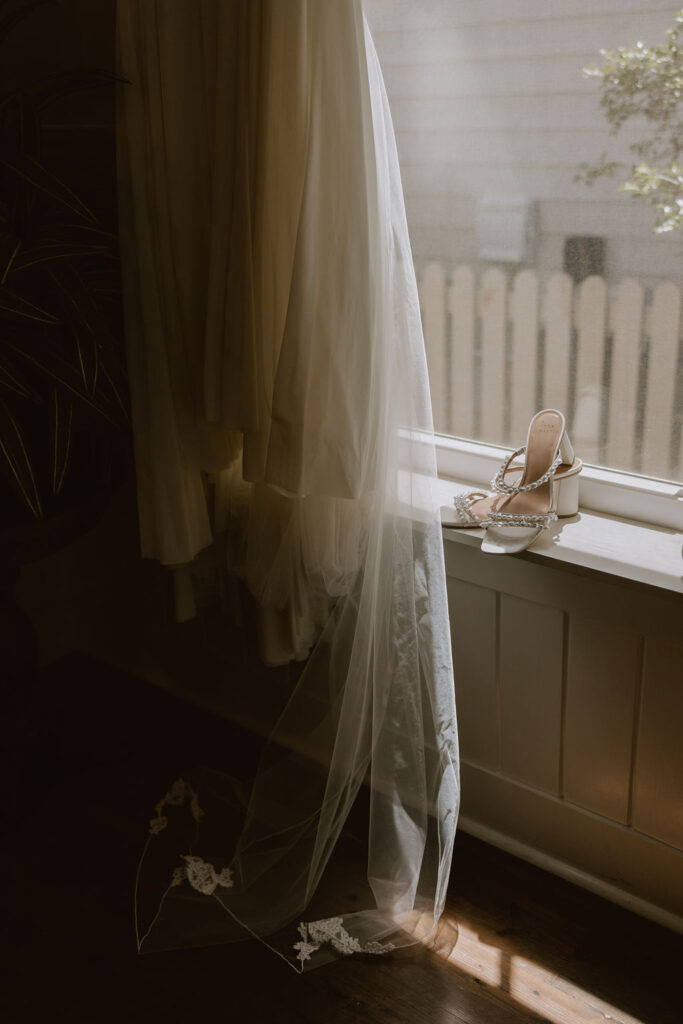 The brides heels sit on a windowsill next to her hanging wedding dress